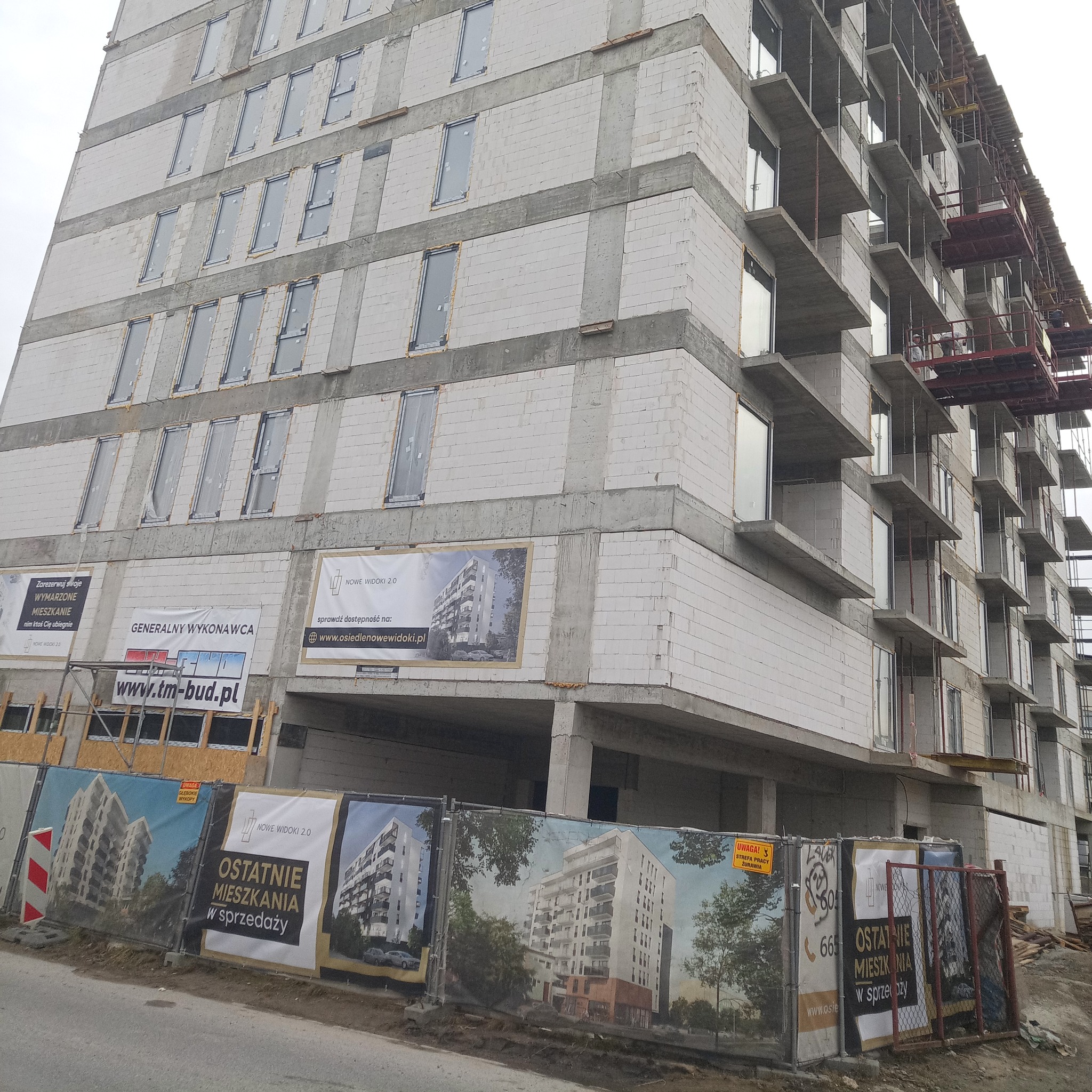 Wielopiętrowy budynek w stanie surowym, z widocznymi pustakami i szalunkami balkonowymi, zabezpieczony siatką z reklamami dewelopera.