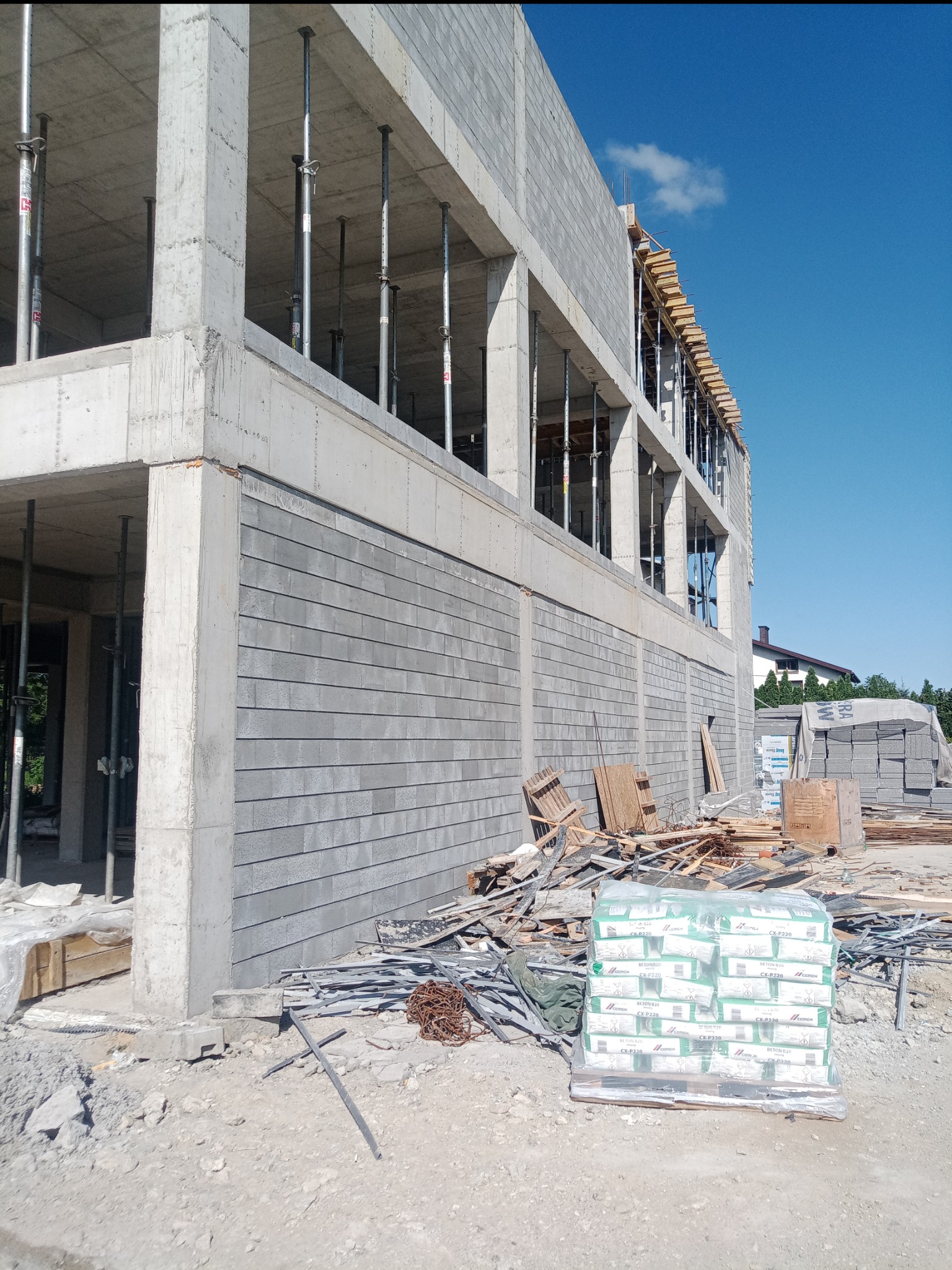 Wznoszony budynek z betonu komórkowego, widoczne podpory stropu, materiały budowlane na placu budowy, błękitne niebo w tle.