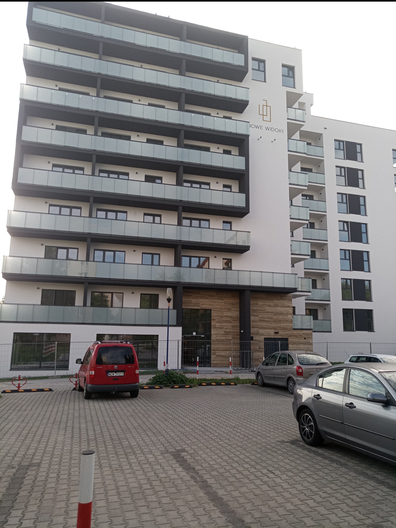 Nowoczesny, wielopiętrowy budynek mieszkalny z balkonami, o białej elewacji z ciemnymi akcentami, widoczny z poziomu parkingu z zaparkowanymi samochodami i logo 'NOWE WIDOKI' na fasadzie.