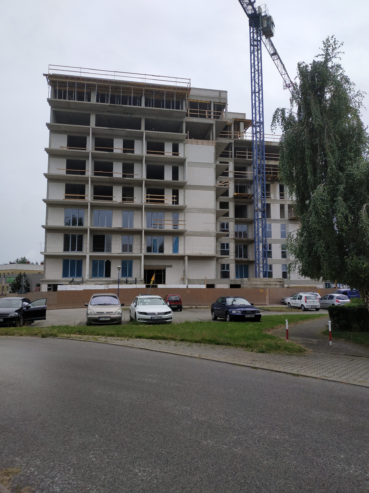 Wielopiętrowy budynek w stanie surowym zamkniętym z oknami, z niebieskim dźwigiem budowlanym obok, na pierwszym planie zaparkowane samochody.