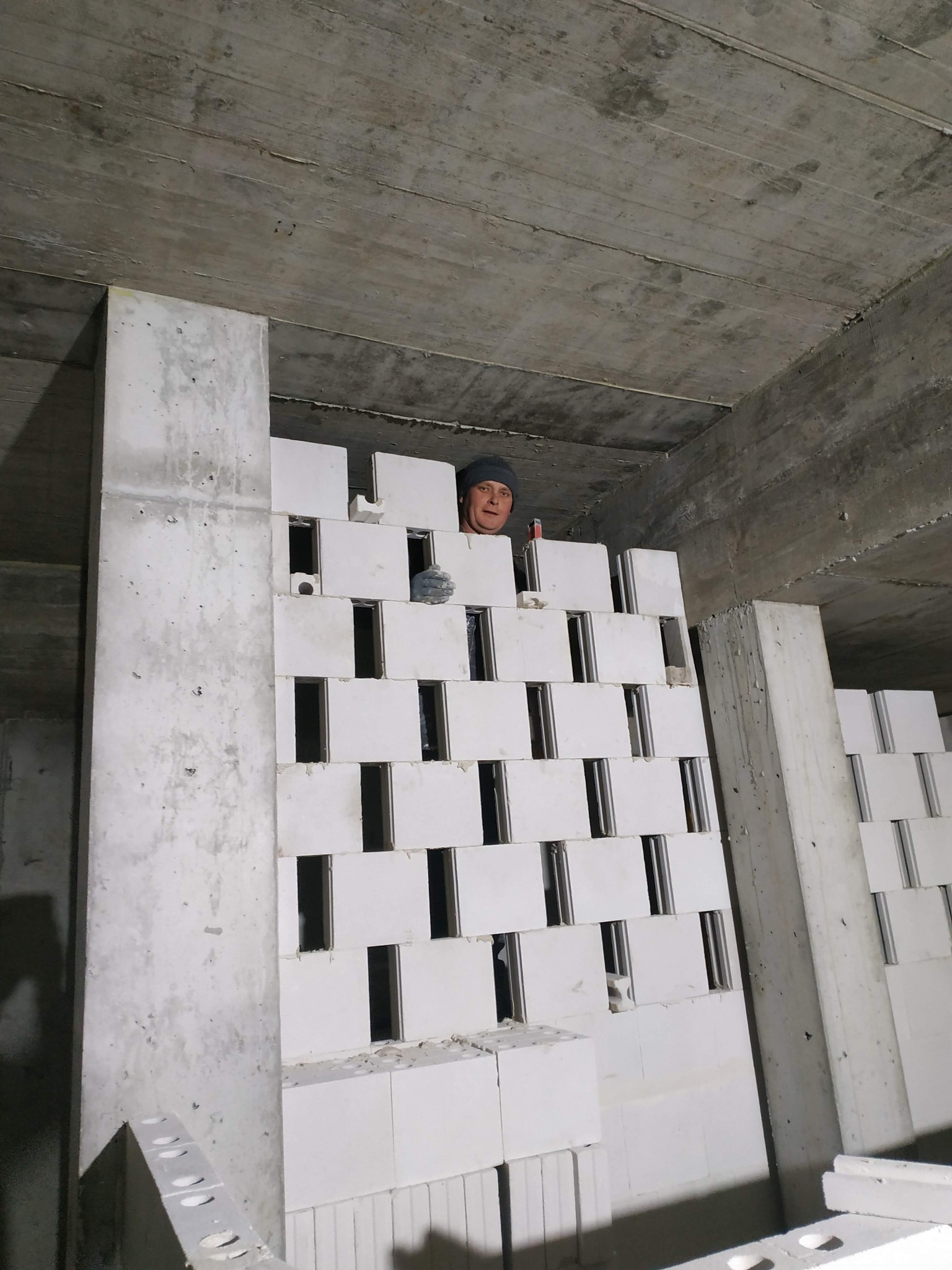 Wznoszenie ściany z betonu komórkowego Ytong w surowym stanie, widoczny murarz w czapce i rękawicy, betonowy strop i filar