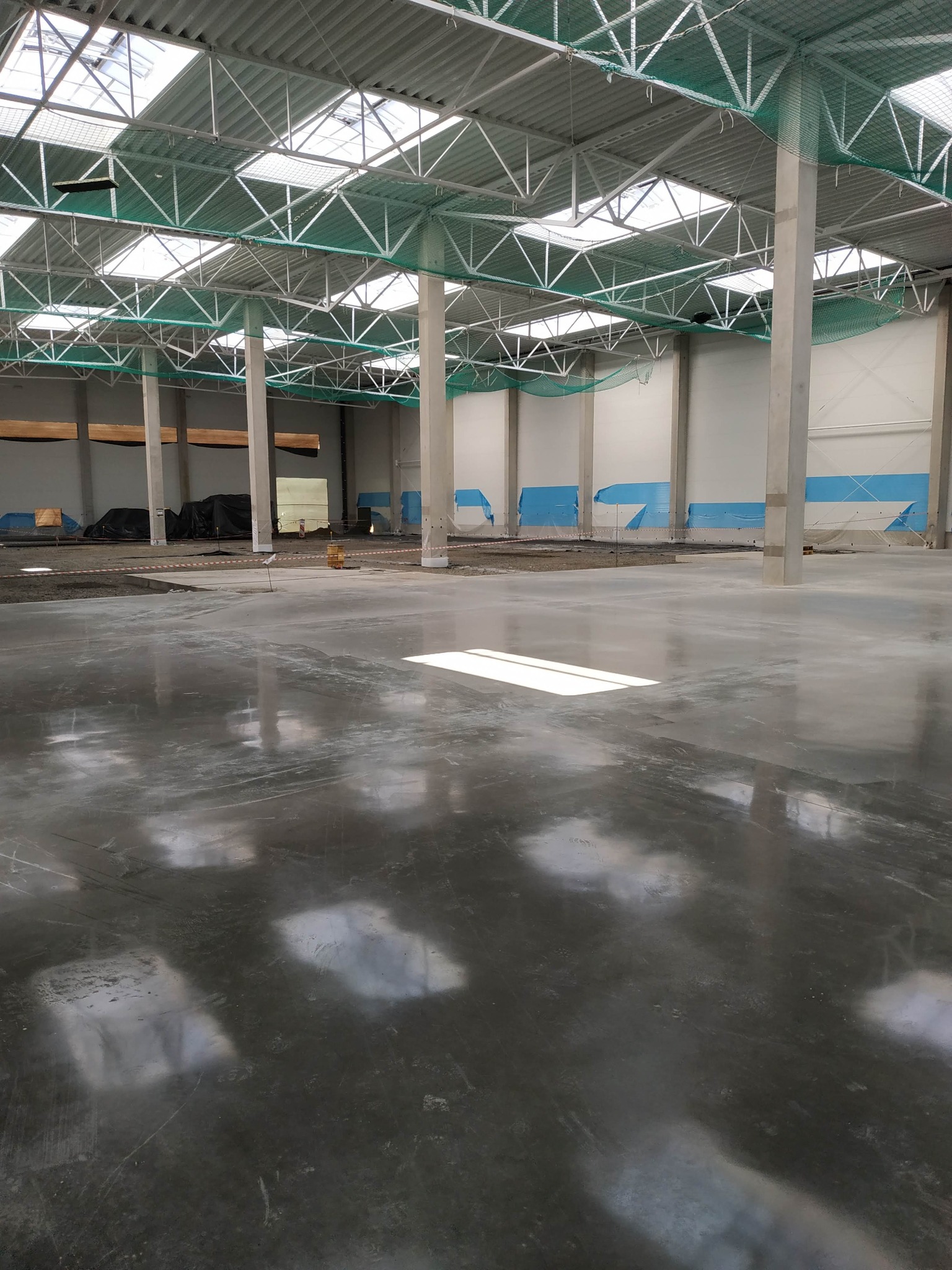 Wnętrze hali produkcyjnej z betonową posadzką w trakcie budowy, widoczne stalowe słupy konstrukcyjne, zabezpieczone ściany, oraz fragmenty dachu z oknami doświetlającymi.