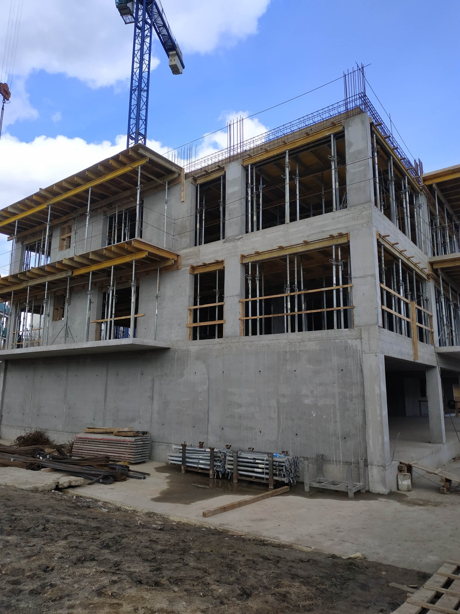 Surowy budynek żelbetowy w trakcie budowy, widoczne szalunki, zbrojenia i rusztowania, na pierwszym planie materiały budowlane i teren budowy, w tle niebieski dźwig i błękitne niebo.
