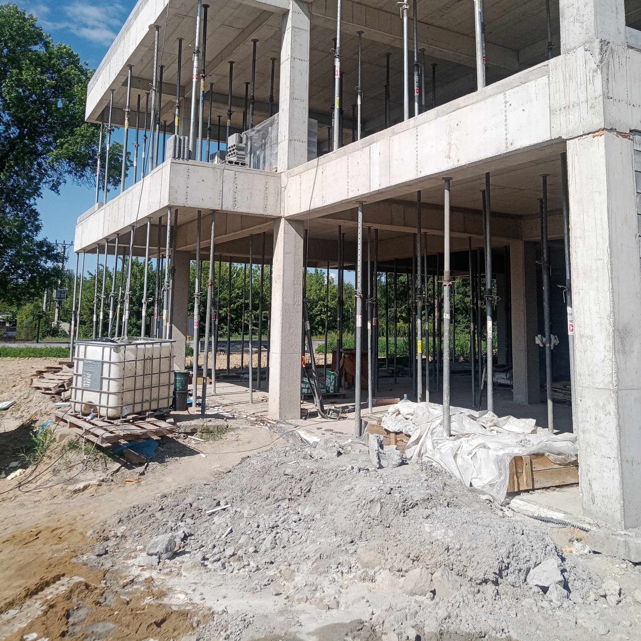 Surowa konstrukcja żelbetowa budynku w trakcie budowy, z widocznymi podporami i szalunkami, na tle zieleni drzew.