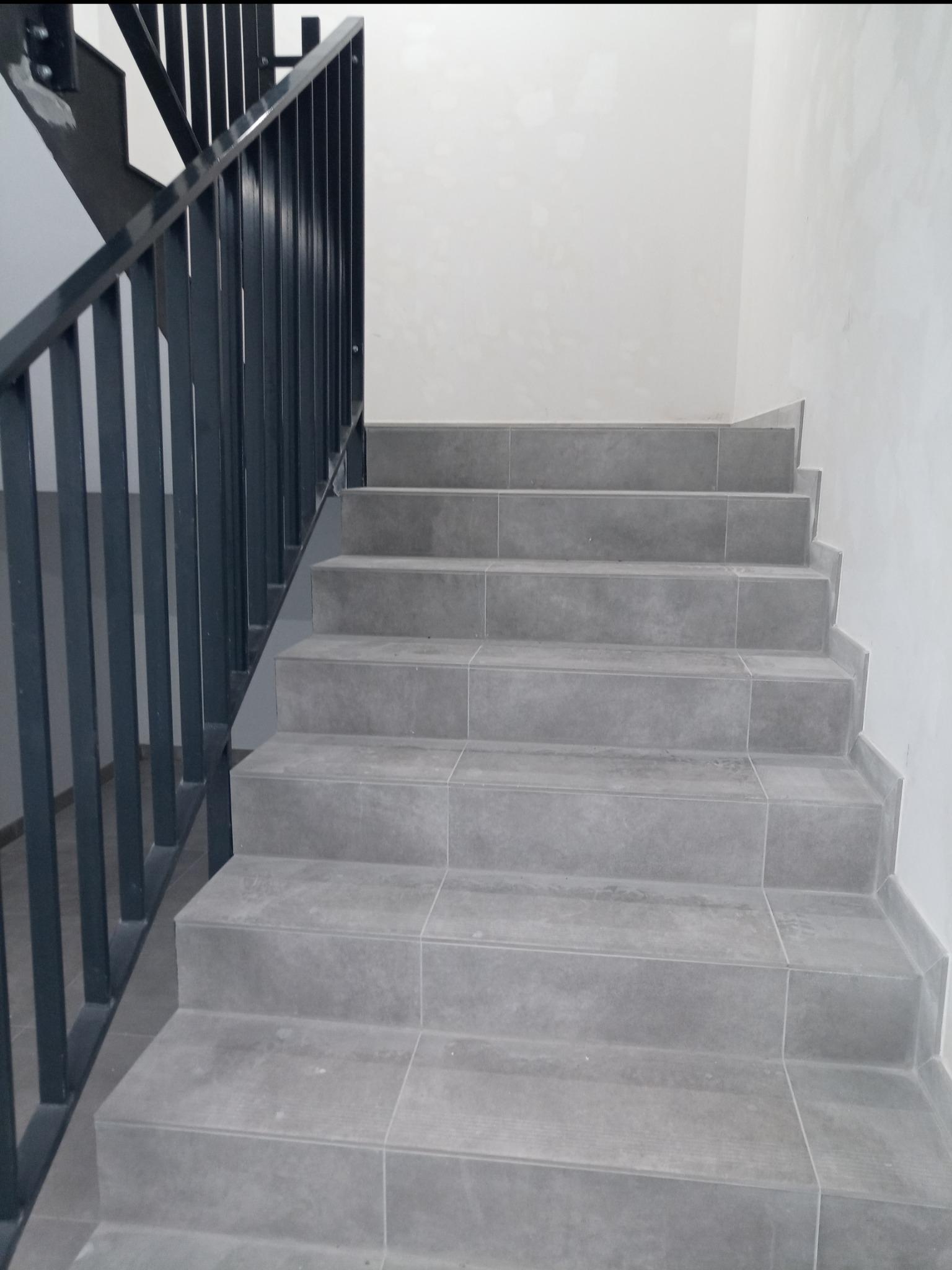 Schody wewnętrzne wykończone szarymi płytkami, z czarną metalową balustradą, widok z dołu ku górze.