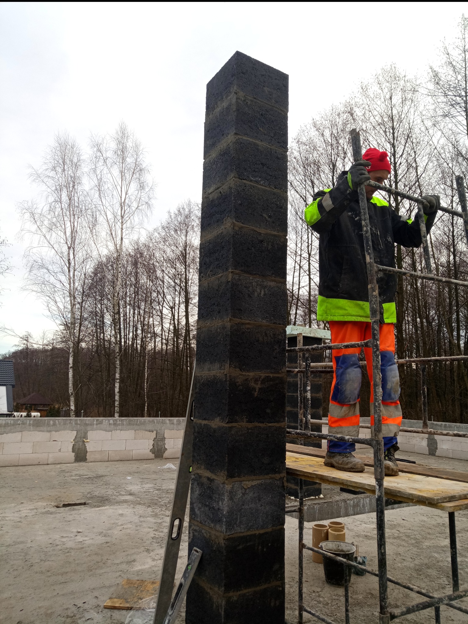 Murarz na rusztowaniu w trakcie wznoszenia komina z ciemnych bloczków na tle surowego stanu budynku i drzew bez liści.