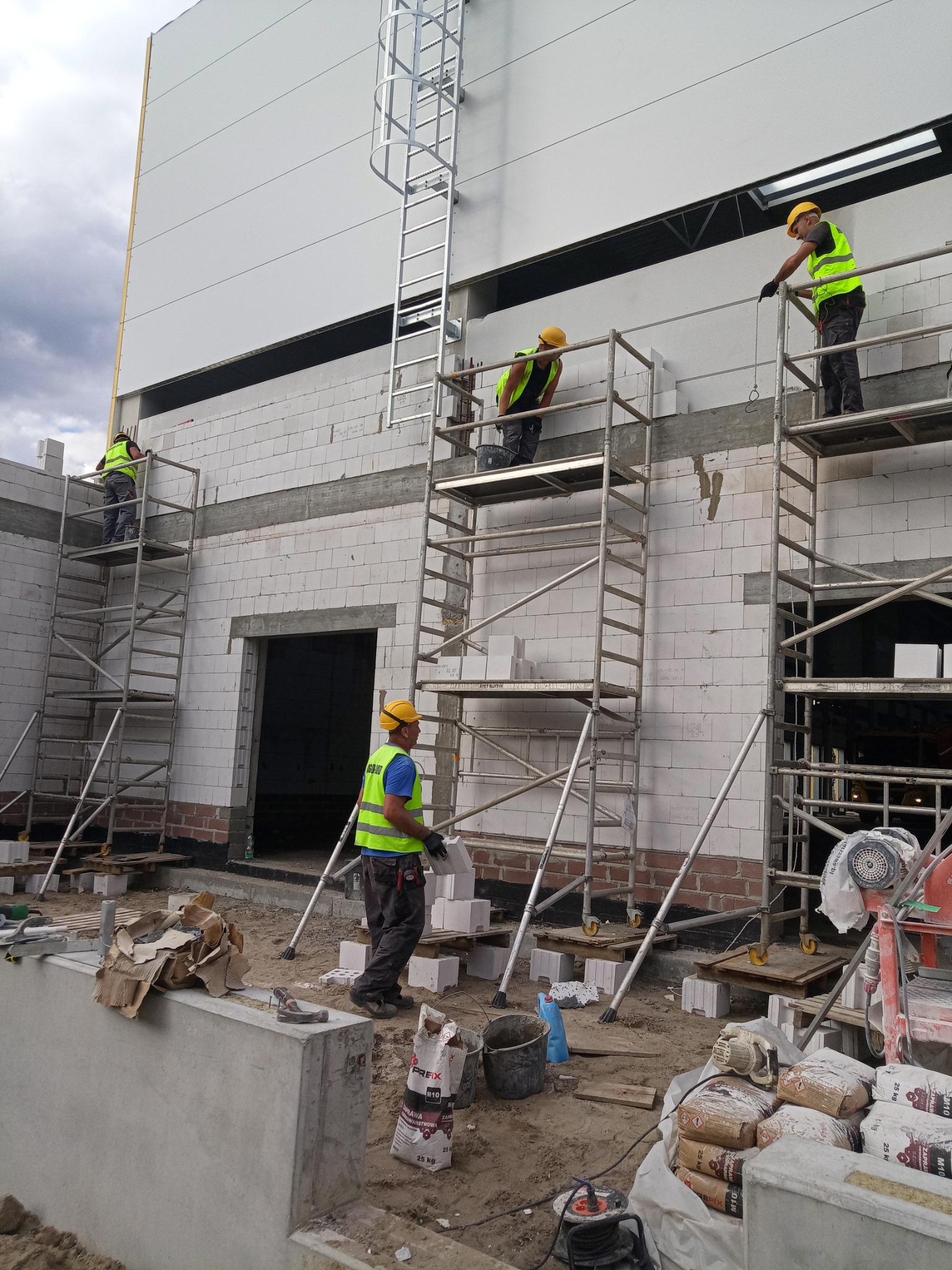 Czterech murarzy w żółtych kaskach i odblaskowych kamizelkach na rusztowaniach podczas wznoszenia ściany z białych bloczków betonu komórkowego Ytong; widoczna drabina przymocowana do ściany...