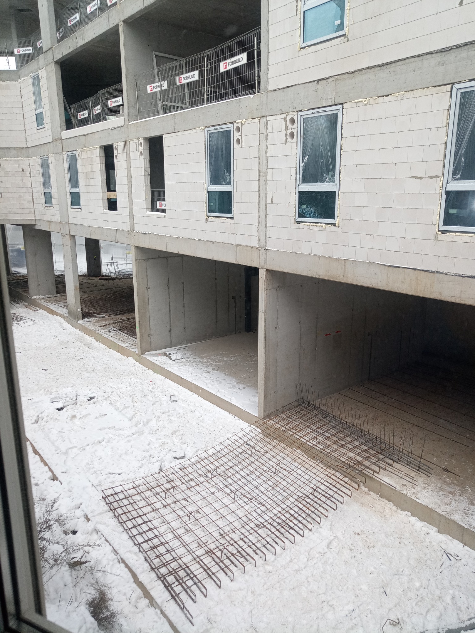 Wnętrze w surowym stanie budynku żelbetowego w trakcie budowy zimą, widoczne zbrojenie na śniegu, ściany z betonu komórkowego i okna zabezpieczone folią.