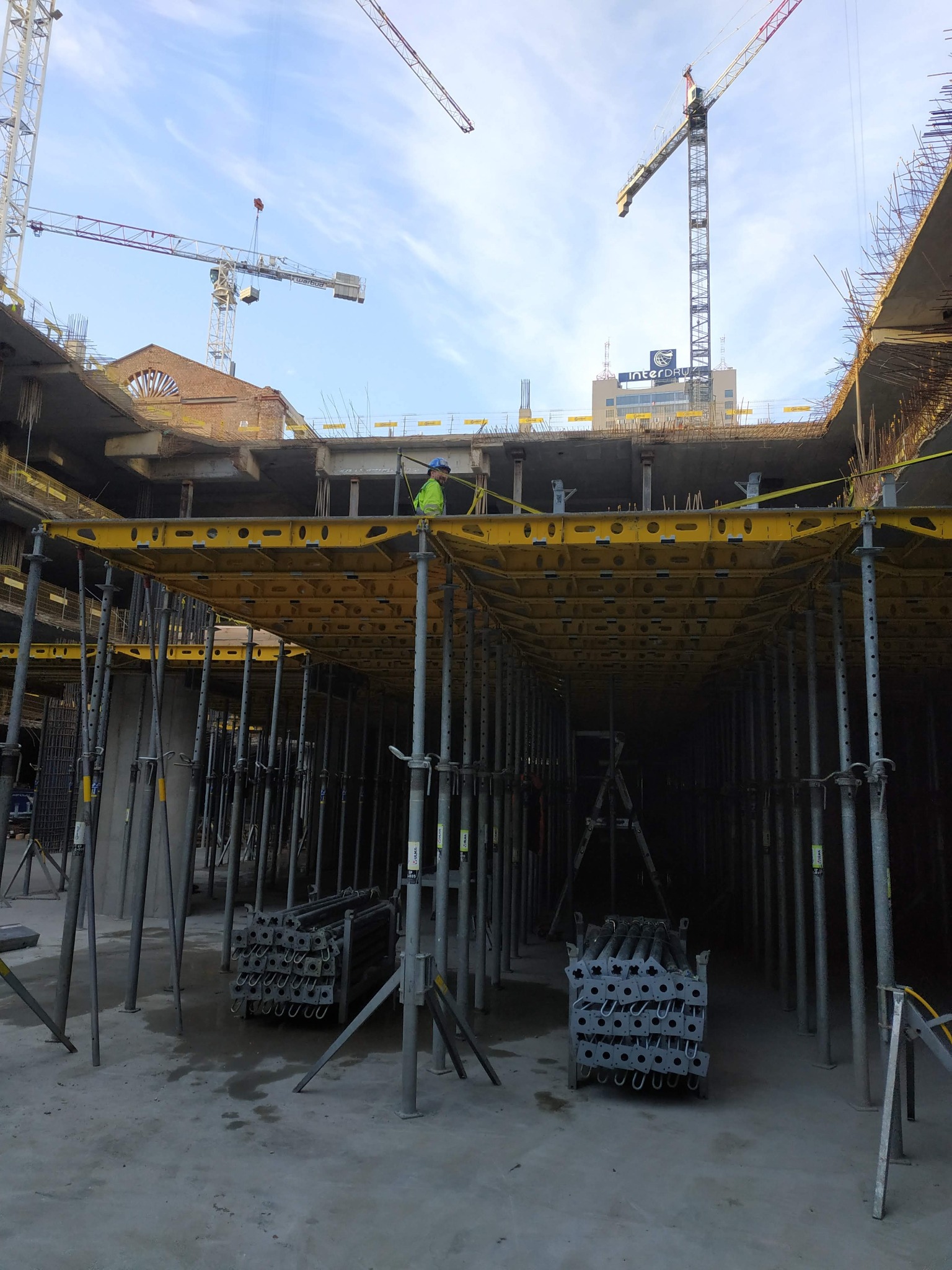 Wnętrze budowy z widoczną konstrukcją szalunkową pod przyszły strop, podpory stalowe, pracownik w odblaskowej kamizelce i żurawie budowlane w tle.
