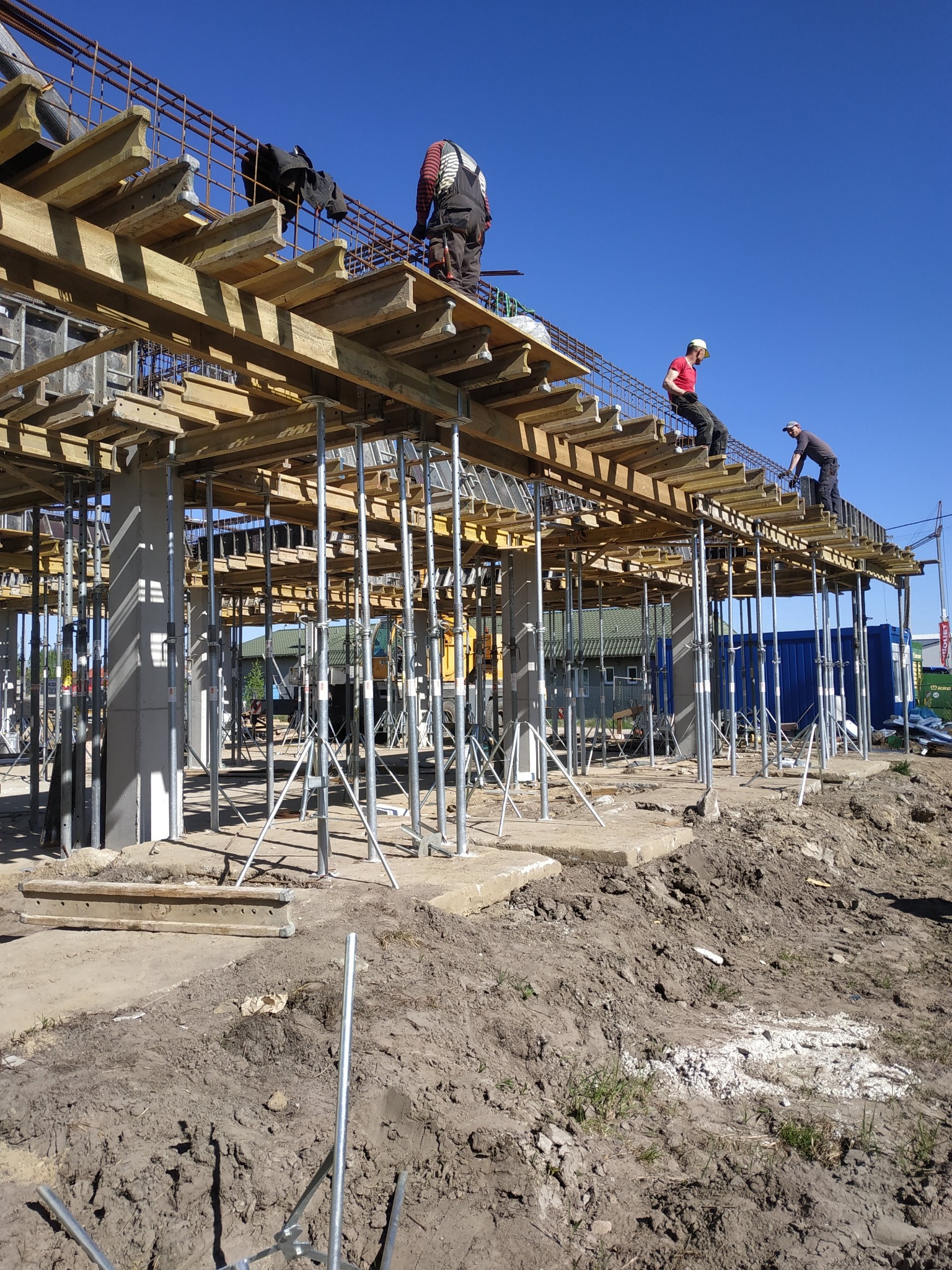 Trzech robotników pracuje na drewnianym szalunku zbrojonym stalą, wspartym na metalowych podporach, na placu budowy w słoneczny dzień.