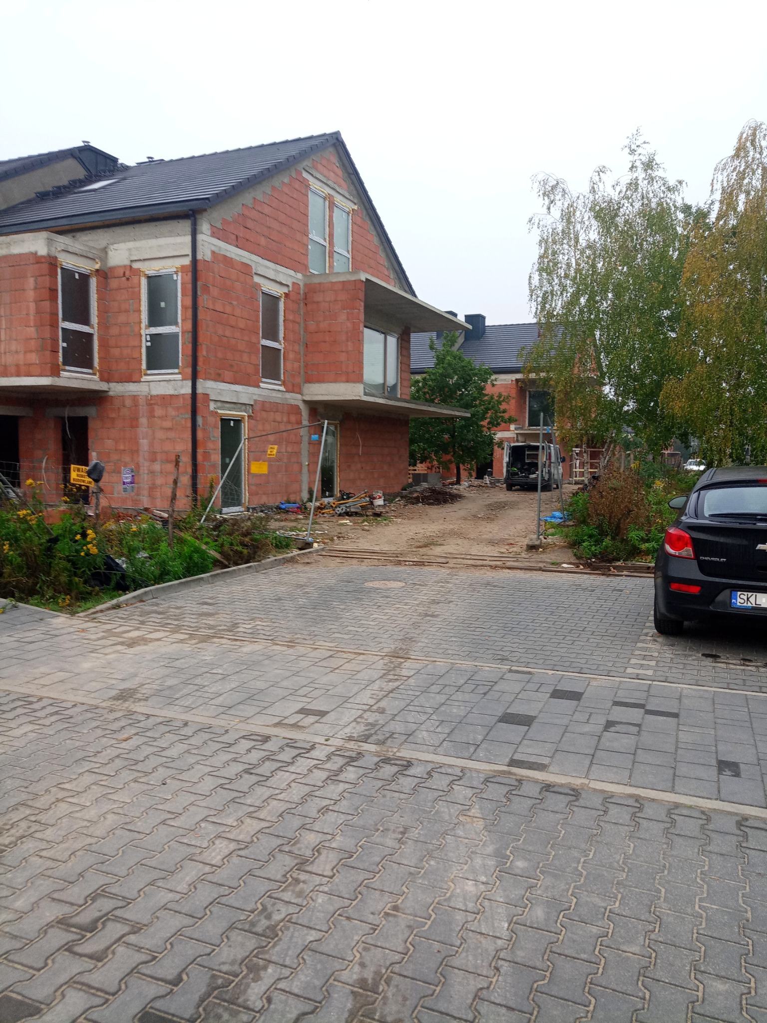 Budowa bliźniaka z czerwonej cegły, widoczne niezadaszone okna, teren budowy z ogrodzeniem i zaparkowany samochód w tle.