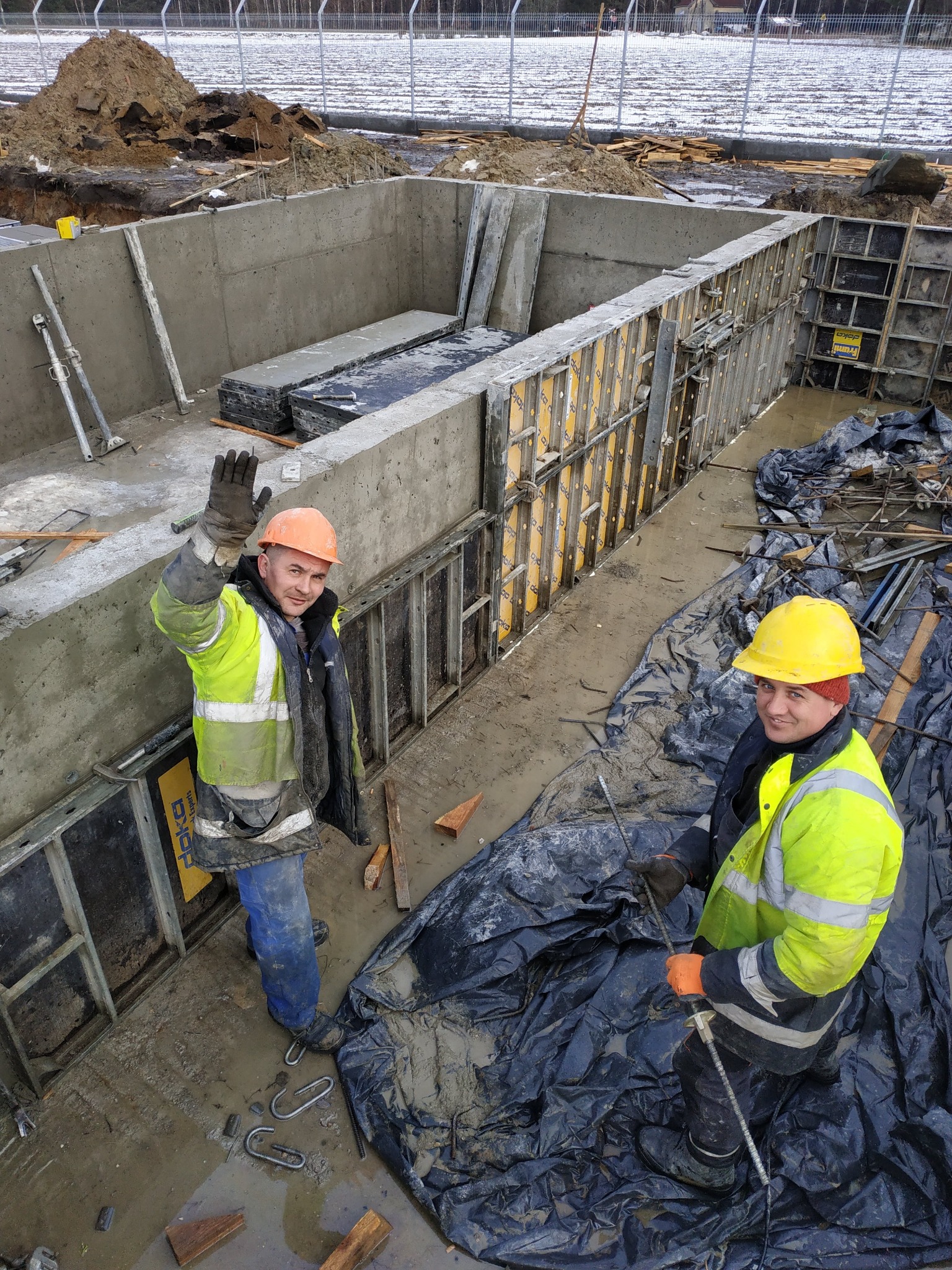 Dwóch robotników w kaskach i odblaskowych kurtkach przy budowie betonowego fundamentu, jeden macha ręką, widoczne szalunki i zbrojenia.