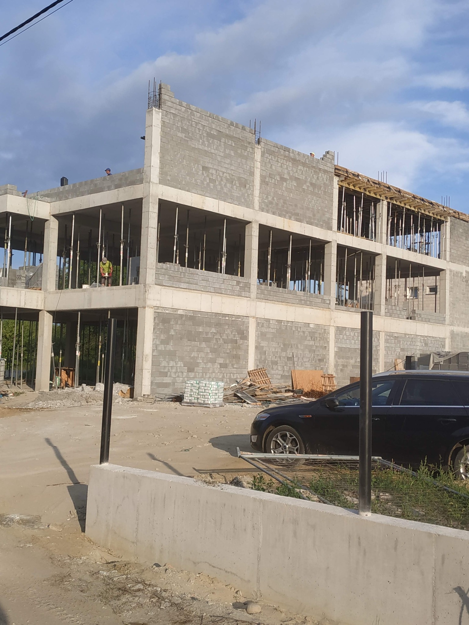 Wznoszenie dwukondygnacyjnego budynku z szarych bloczków, widoczne podpory stropu, zbrojenie, na górze budynku dwóch pracowników, obok zaparkowany czarny samochód osobowy.