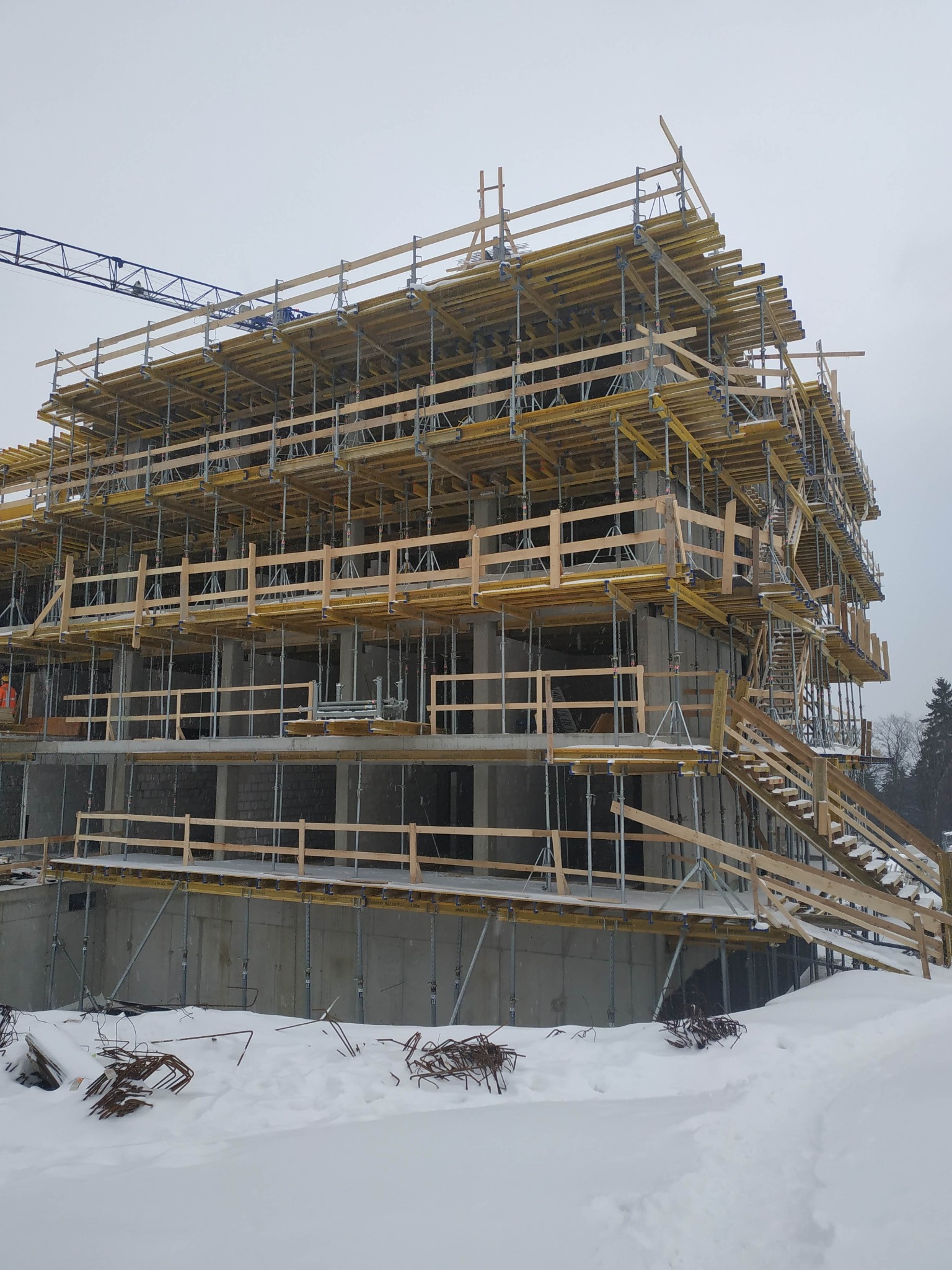 Wielopiętrowy budynek w stanie surowym, otoczony rusztowaniami i zasypany śniegiem, z widocznym żurawiem w tle i zbrojeniami na pierwszym planie.