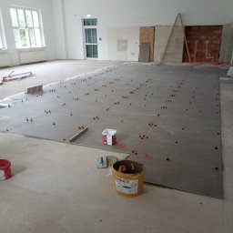 LUKAS BAU ŁUKASZ MŚCICHOWSKI - Układanie szarych płytek na podłodze z użyciem systemu poziomowania, widoczne narzędzia glazurnicze i wiadro z klejem. Duże, jasne pomieszczenie w trakcie prac wykończeniowych.