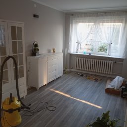 LUKAS BAU ŁUKASZ MŚCICHOWSKI - Jasne wnętrze po remoncie: szare panele, białe meble, okno z firankami. Widoczny odkurzacz i narzędzia, co sugeruje etap sprzątania po pracach wykończeniowych.