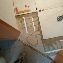 Montaż ogrzewania podłogowego w łazience, widoczne kable grzewcze, płytki w trakcie układania, poziomica i elementy mocujące. Prace wykończeniowe w toku.