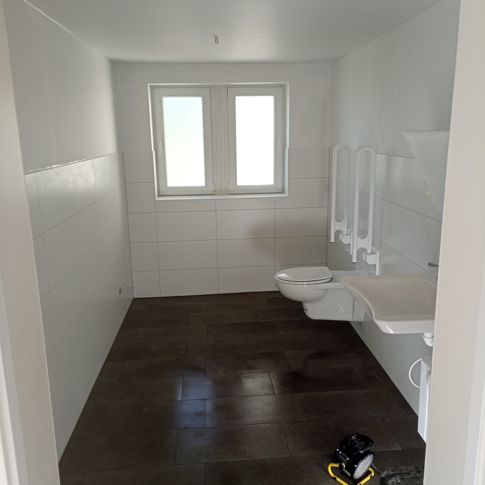 Biała łazienka z brązowymi płytkami podłogowymi. Umywalka i toaleta zamontowane na ścianie. Uchwyty dla niepełnosprawnych. Widoczny wentylator pod umywalką.