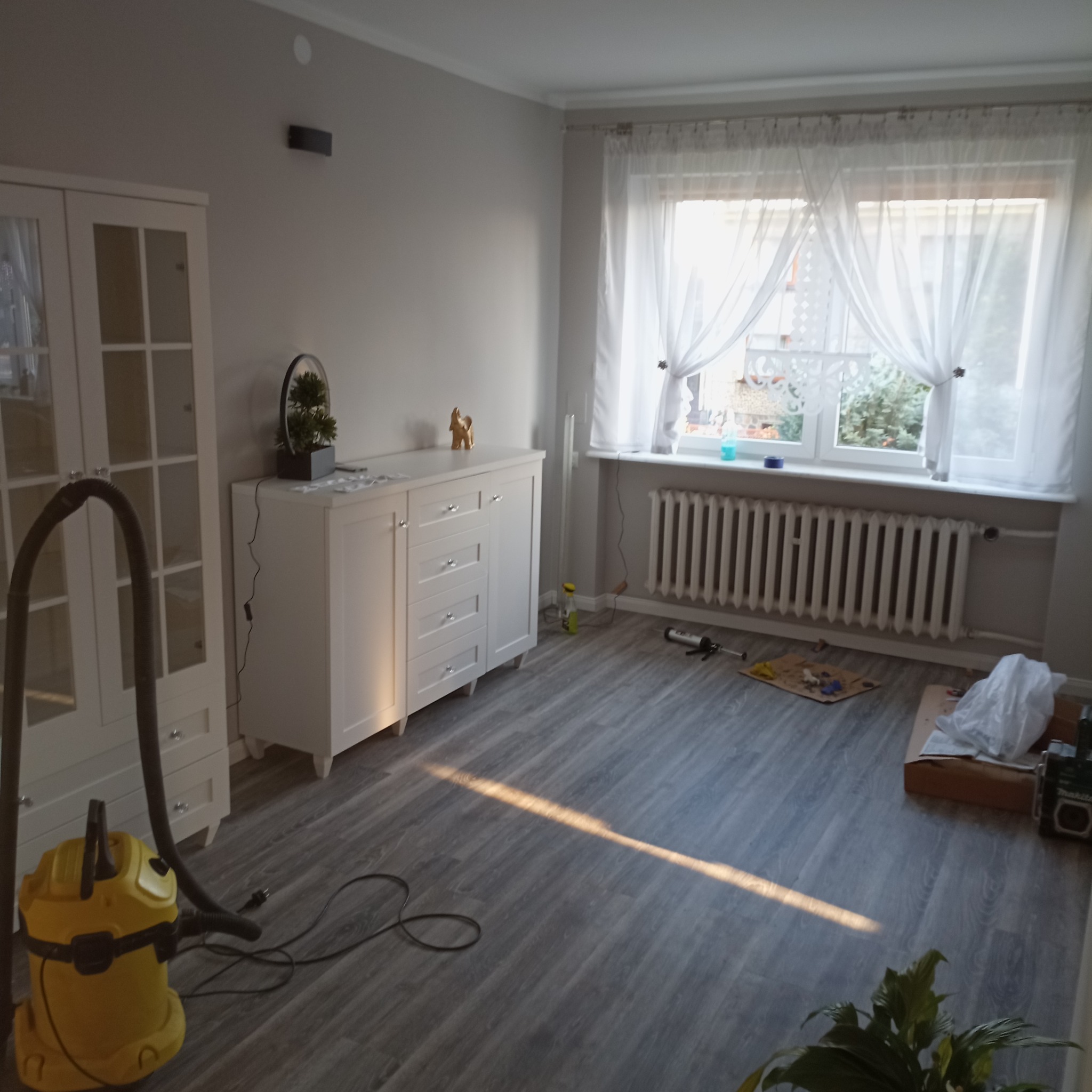 Jasne wnętrze po remoncie: szare panele, białe meble, okno z firankami. Widoczny odkurzacz i narzędzia, co sugeruje etap sprzątania po pracach wykończeniowych.