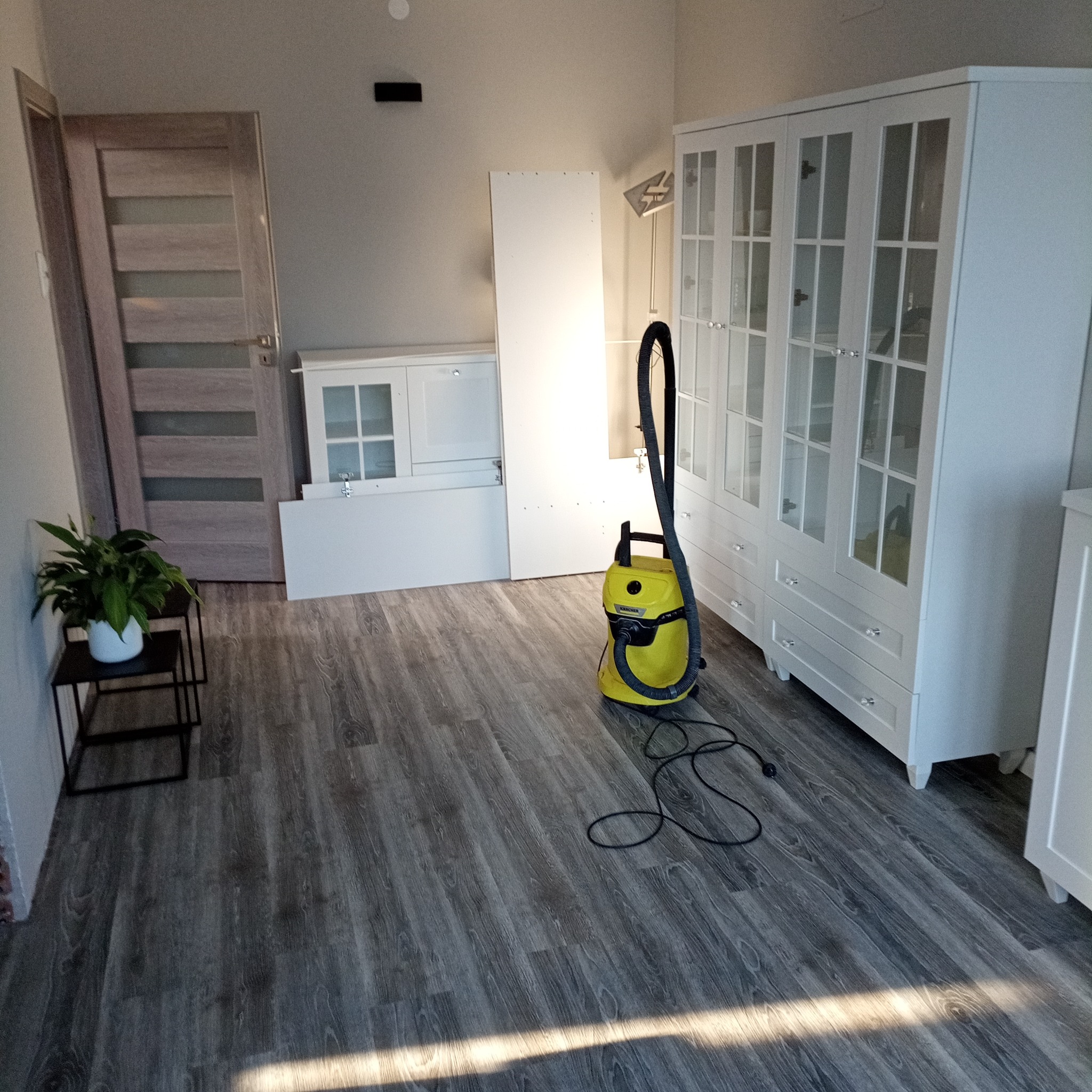Pomieszczenie po remoncie z odkurzaczem Karcher, nowa podłoga, białe meble w trakcie montażu i doniczka z rośliną.