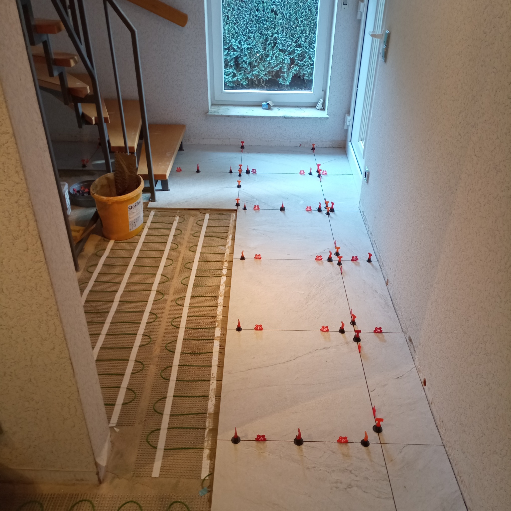 Układanie jasnych płytek podłogowych z systemem poziomowania, widoczny fragment instalacji ogrzewania podłogowego i schody. Prace wykończeniowe w korytarzu.