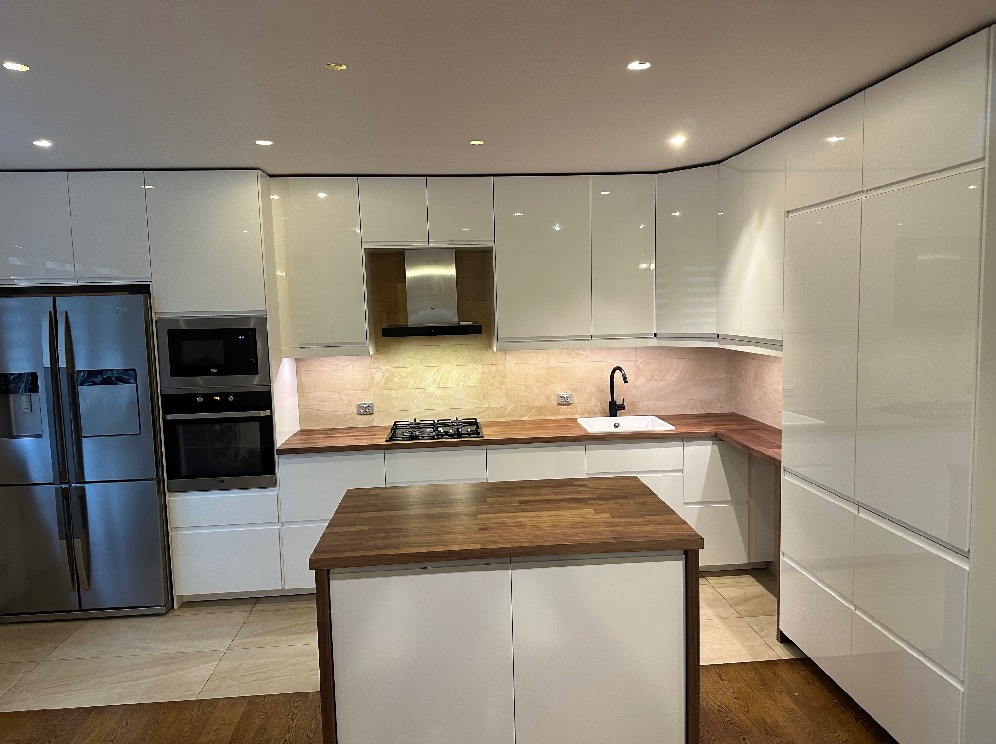 Nowoczesna kuchnia z białymi, lakierowanymi frontami szafek, drewnianym blatem i wyspą kuchenną z ciemnym blatem, oświetleniem punktowym i sprzętem AGD.