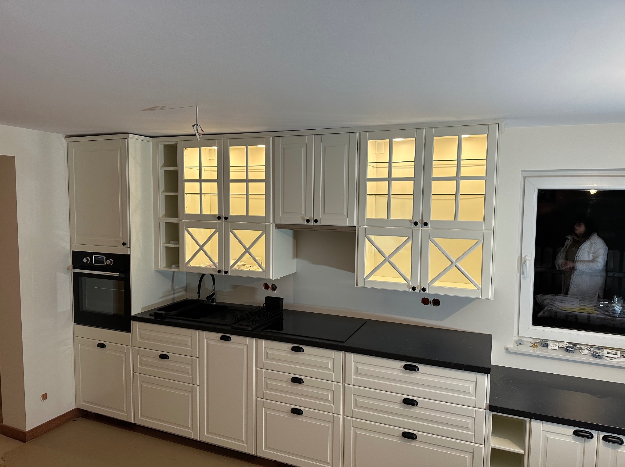Nowoczesna kuchnia z białymi szafkami, czarnym blatem i oświetleniem LED w górnych szafkach z przeszklonymi frontami, widoczne niedokończone prace elektryczne.