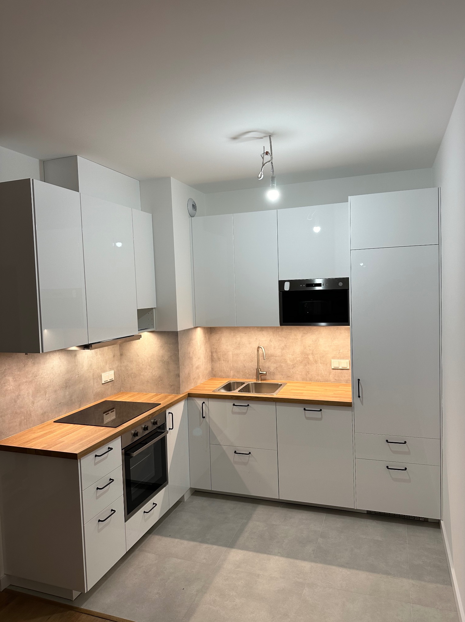 Nowoczesna, jasna kuchnia z białymi, lakierowanymi szafkami i drewnianymi blatami, oświetlona ciepłym światłem spod szafek wiszących.