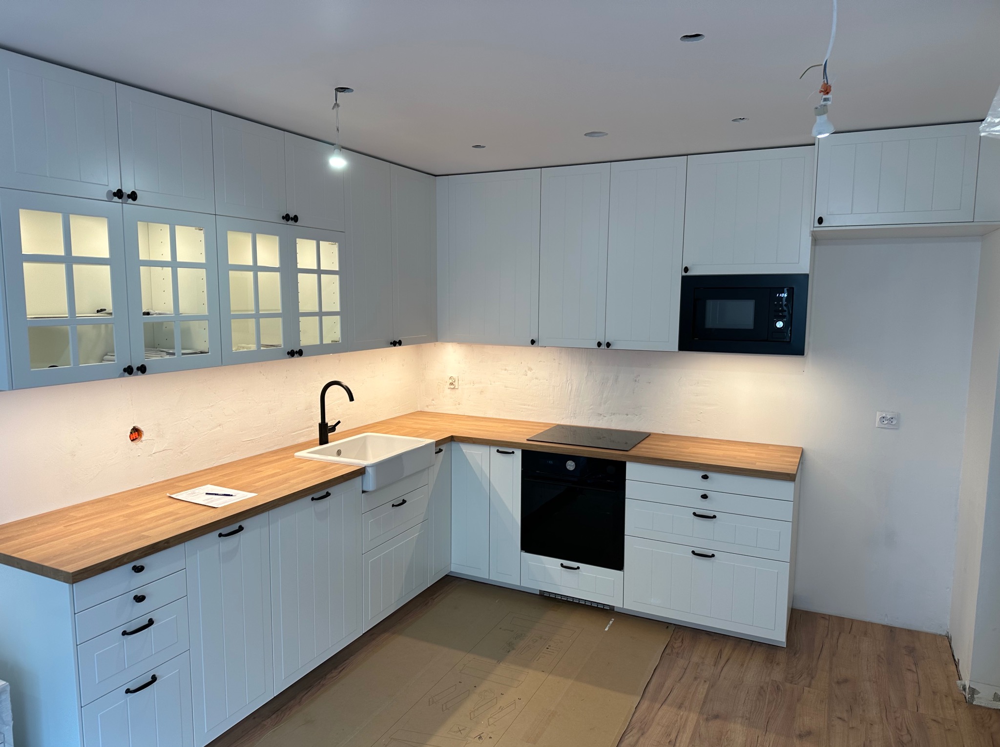 Nowoczesna, biała kuchnia z drewnianym blatem, czarną armaturą i sprzętem AGD, w trakcie wykańczania, z widocznymi niedokończonymi elementami instalacji elektrycznej i kartonem ochronnym na podłodze.