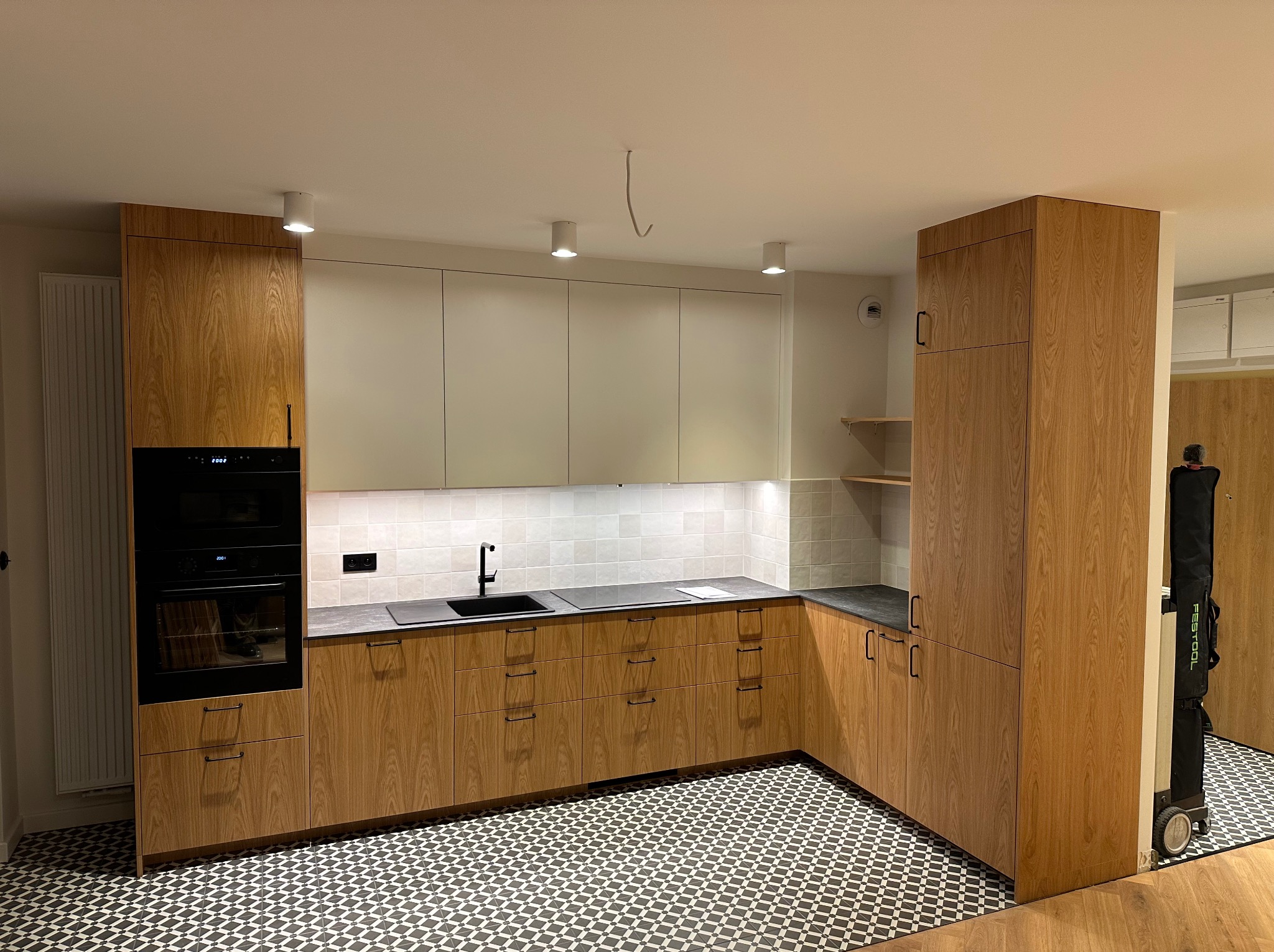 Nowoczesna kuchnia z drewnianymi frontami szafek, czarnym blatem i geometryczną mozaiką na podłodze. Widoczny piekarnik w zabudowie i czarna bateria zlewozmywakowa.