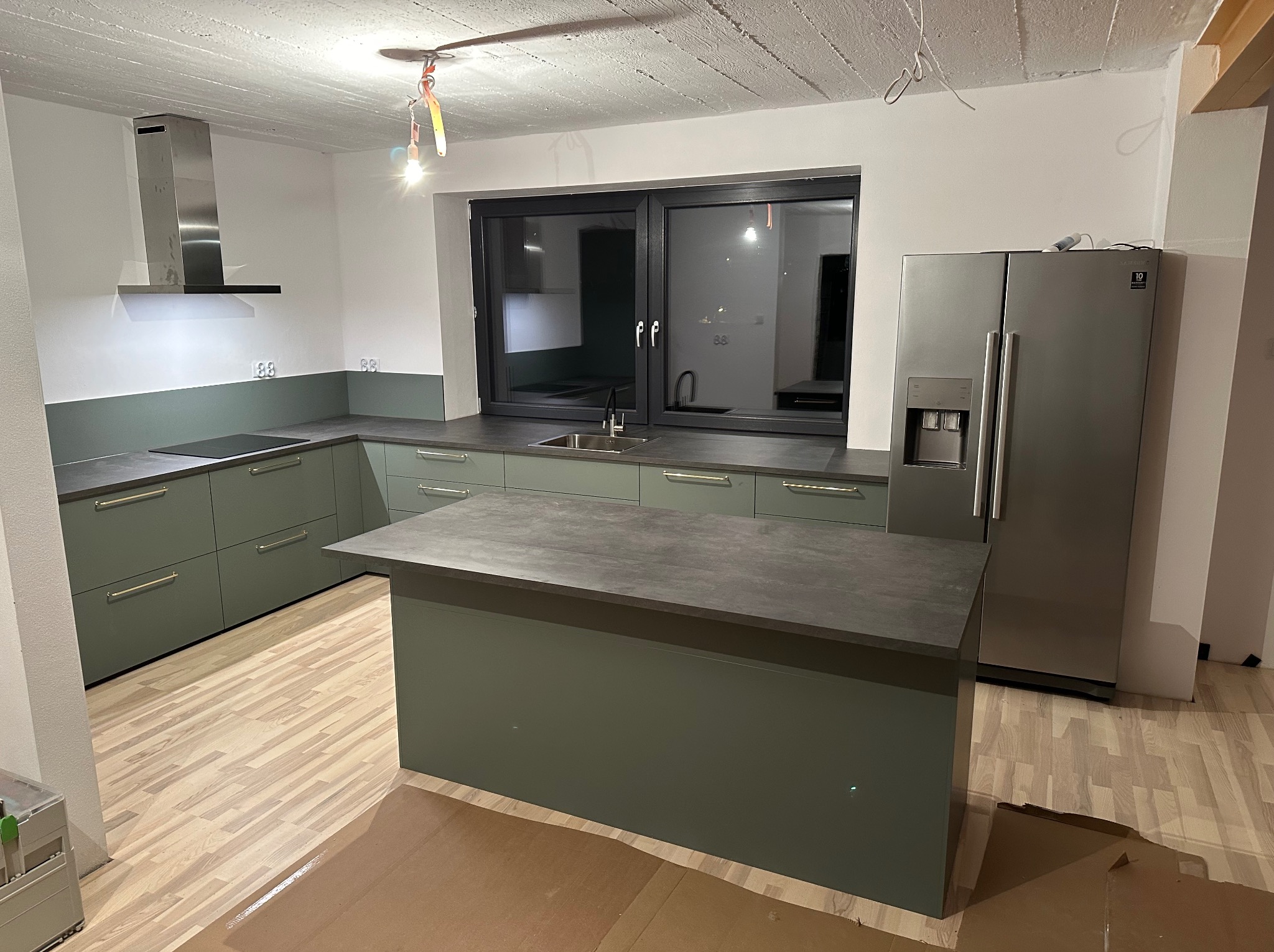 Nowoczesna kuchnia z zielonymi szafkami i wyspą, surowy betonowy sufit z widocznymi kablami, podłoga częściowo zabezpieczona kartonem.