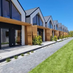 Radosław Sawicki - Szereg nowoczesnych domków jednorodzinnych z szarymi dachami i drewnianymi pergolami, widok z poziomu chodnika z kostki brukowej i trawnika.