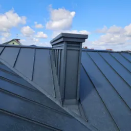 Nowy, grafitowy dach z blachy trapezowej z kominem na tle błękitnego nieba z chmurami.