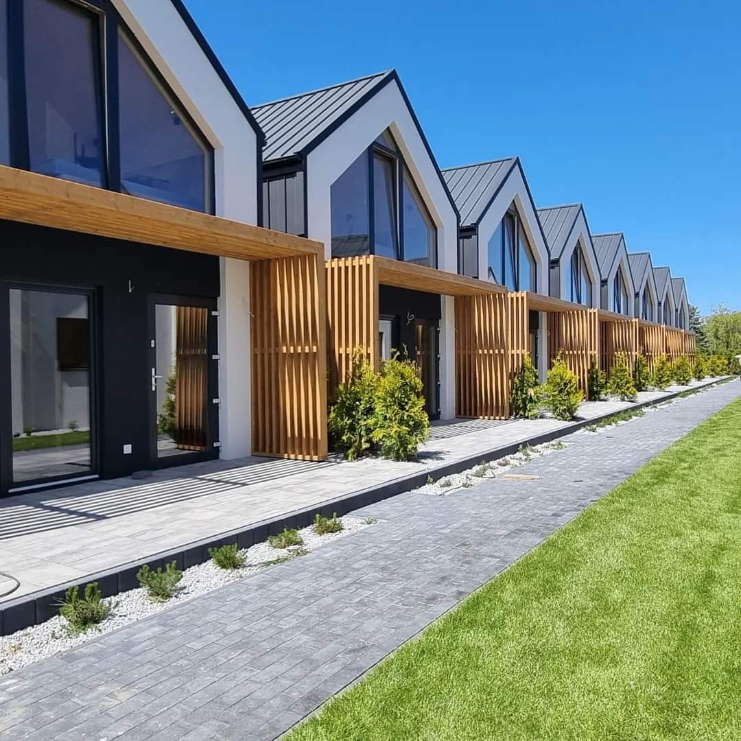 Szereg nowoczesnych domków jednorodzinnych z szarymi dachami i drewnianymi pergolami, widok z poziomu chodnika z kostki brukowej i trawnika.