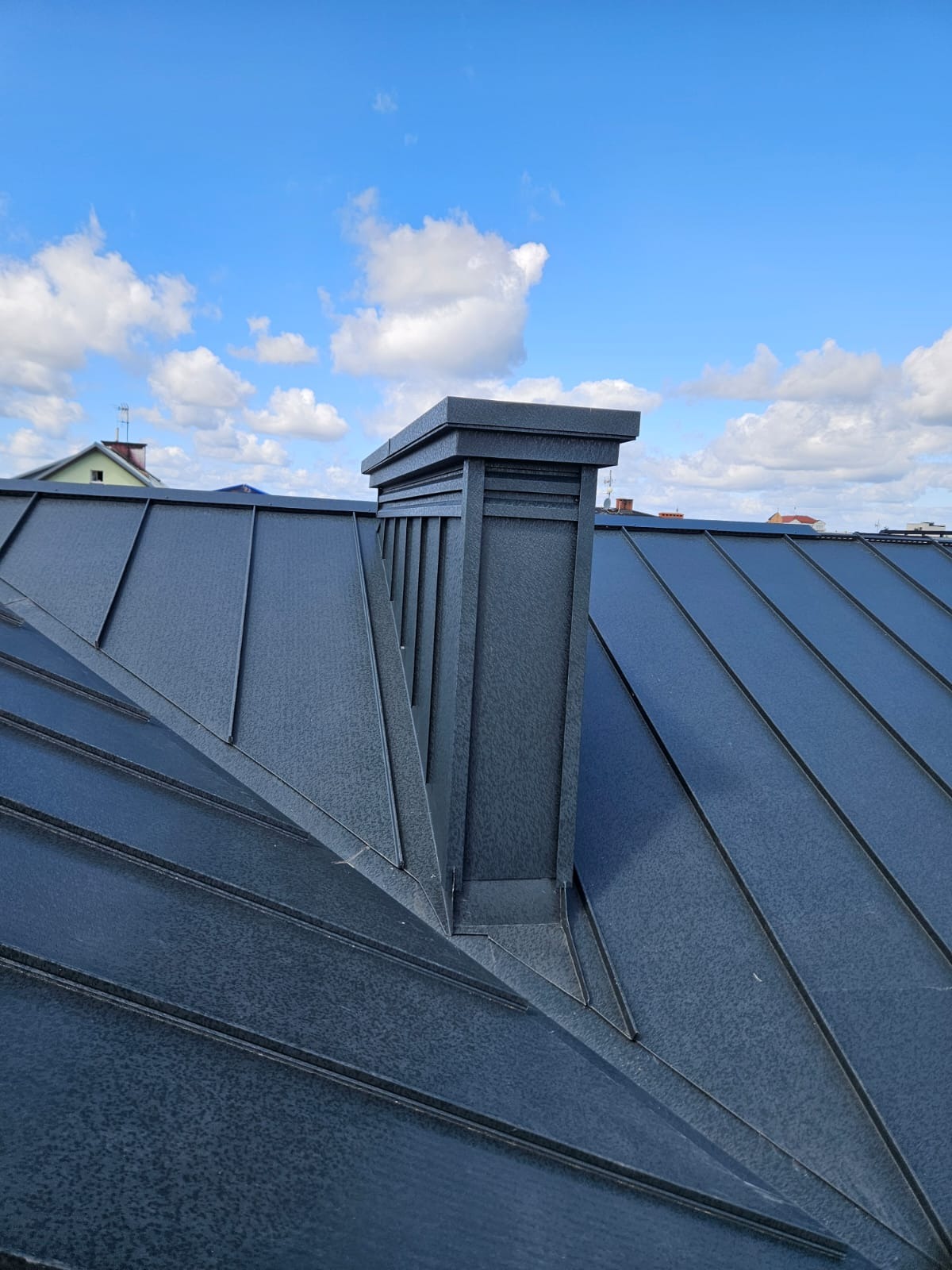 Nowy, grafitowy dach z blachy trapezowej z kominem na tle błękitnego nieba z chmurami.