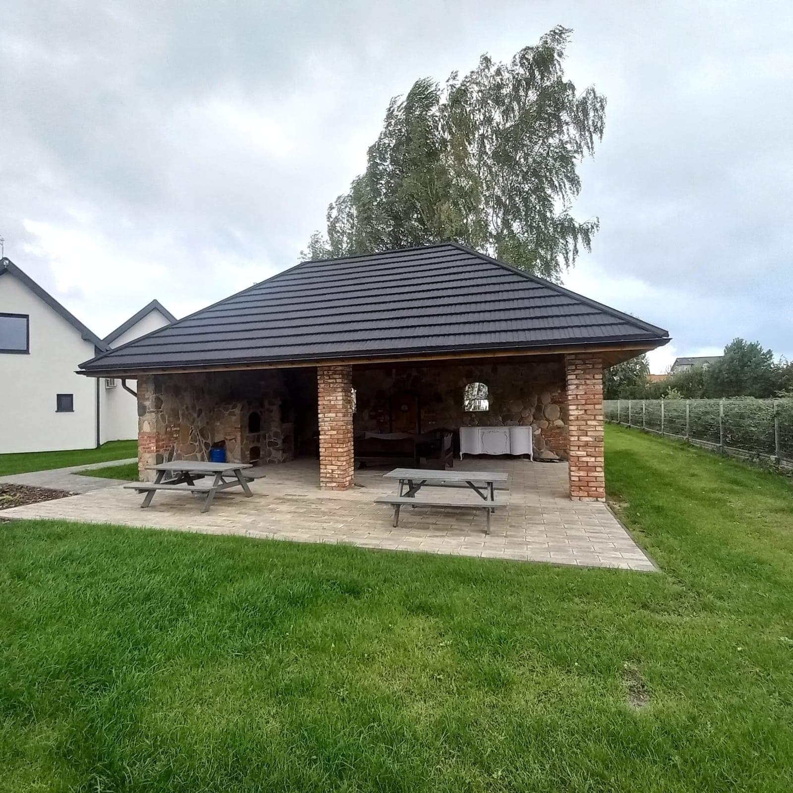 Zadaszona altana z kamienno-ceglanymi ścianami, pokryta ciemną dachówką, z dwoma drewnianymi stołami piknikowymi na zewnątrz, oraz meblami wewnątrz, na tle zielonego trawnika i drzew.