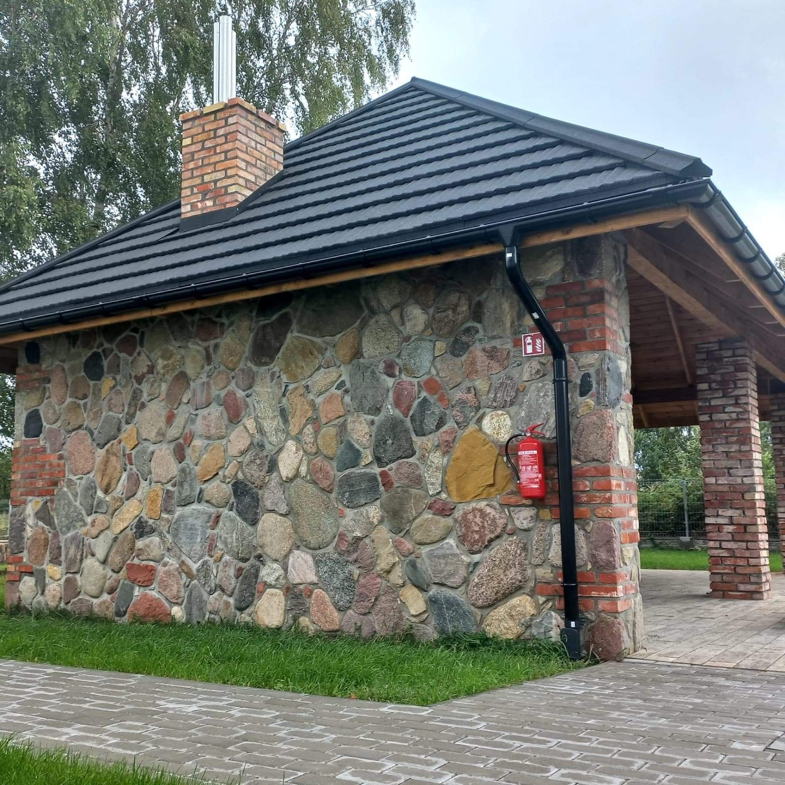 Murowana altana z kamienną elewacją, czarną dachówką i ceglanym kominem, wyposażona w rynnę i gaśnicę.