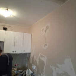 A.P. SOLID ANDRZEJ PAWLAK - Fragment remontowanego wnętrza: ściana z widocznymi śladami szpachlowania, przygotowana do malowania, zakryte folią podłoże, widoczne szafki kuchenne i instalacja elektryczna.