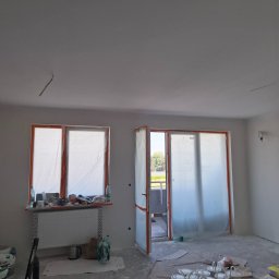 A.P. SOLID ANDRZEJ PAWLAK - Pokój przed malowaniem: okna i drzwi balkonowe zabezpieczone folią, widoczne puszki z farbą na podłodze, ślady po pracach przygotowawczych.