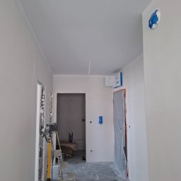 A.P. SOLID ANDRZEJ PAWLAK - Wnętrze w trakcie prac wykończeniowych: świeżo pomalowane ściany na biało, zabezpieczone drzwi folią, widoczna drabina i lampa budowlana, podłoga częściowo zakryta folią malarską, ślady farby...
