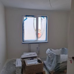 A.P. SOLID ANDRZEJ PAWLAK - Pokój w trakcie remontu, z zabezpieczonym oknem folią malarską, widoczne materiały budowlane na paletach, ślady po pracach na podłodze, kabel wystający z sufitu.