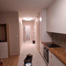 A.P. SOLID ANDRZEJ PAWLAK - Wnętrze mieszkania w trakcie malowania: widoczne puszki z farbą, wiadro, kuweta malarska na podłodze, fragment pomalowanej ściany, zabudowa kuchenna z drewnianym blatem i płytą indukcyjną,...