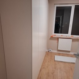 A.P. SOLID ANDRZEJ PAWLAK - Wnętrze pokoju z zamontowaną białą zabudową meblową pod sufit, widoczne okno z grzejnikiem i częściowo zakryte folią zabezpieczającą, na podłodze panele i kartonowe opakowanie. W szybie okna odbija...