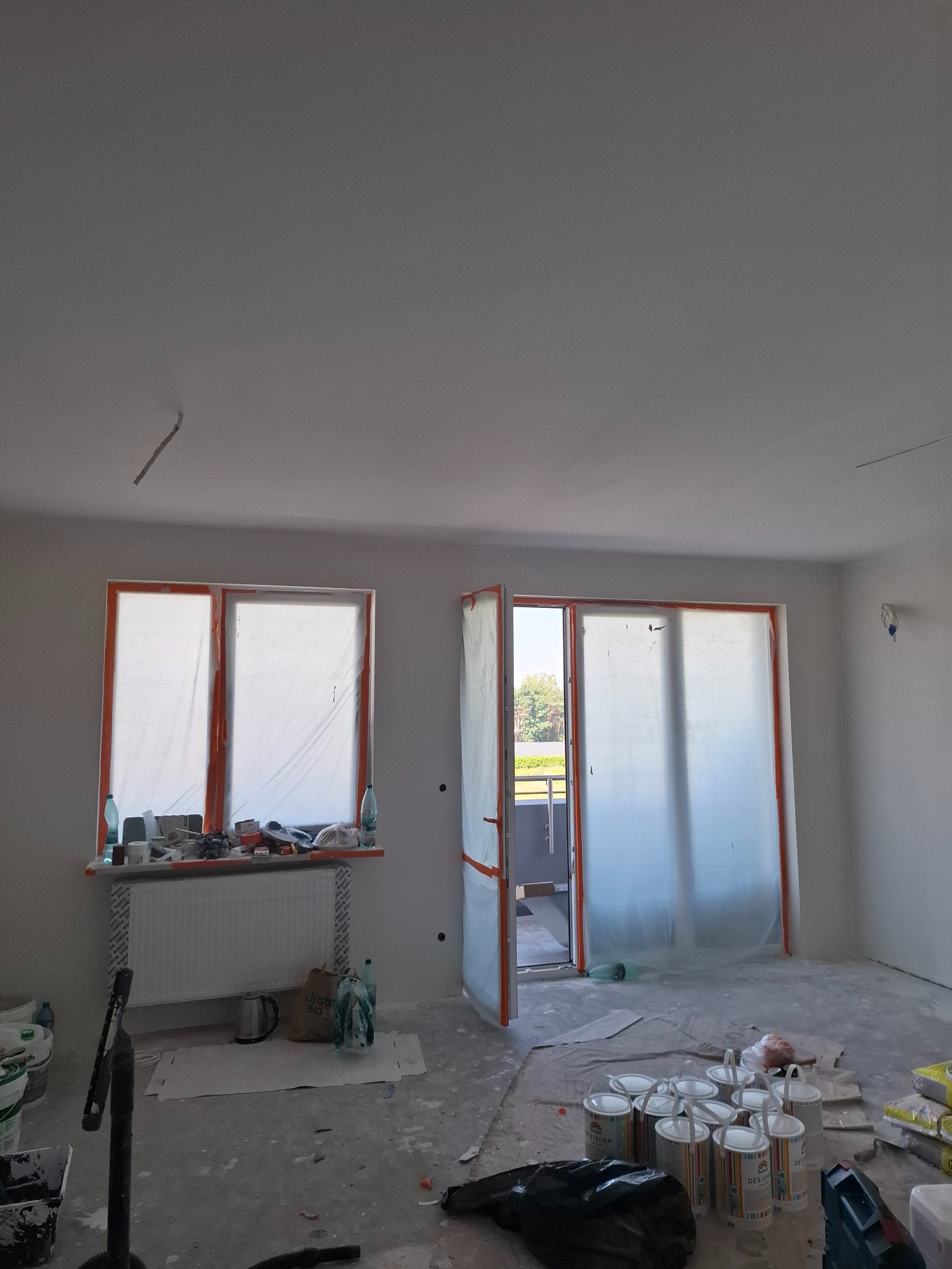 Pokój przed malowaniem: okna i drzwi balkonowe zabezpieczone folią, widoczne puszki z farbą na podłodze, ślady po pracach przygotowawczych.