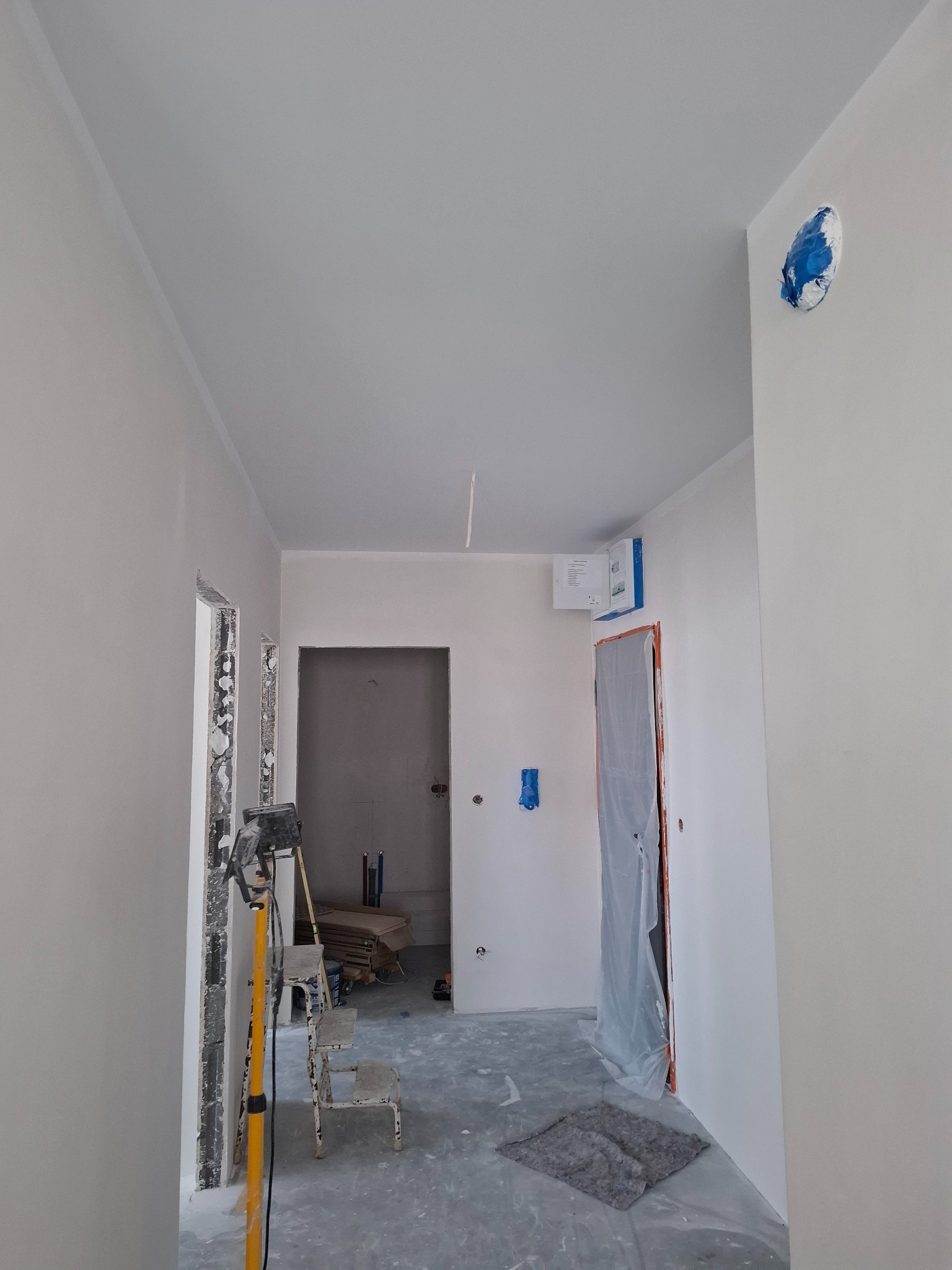 Wnętrze w trakcie prac wykończeniowych: świeżo pomalowane ściany na biało, zabezpieczone drzwi folią, widoczna drabina i lampa budowlana, podłoga częściowo zakryta folią malarską, ślady farby...