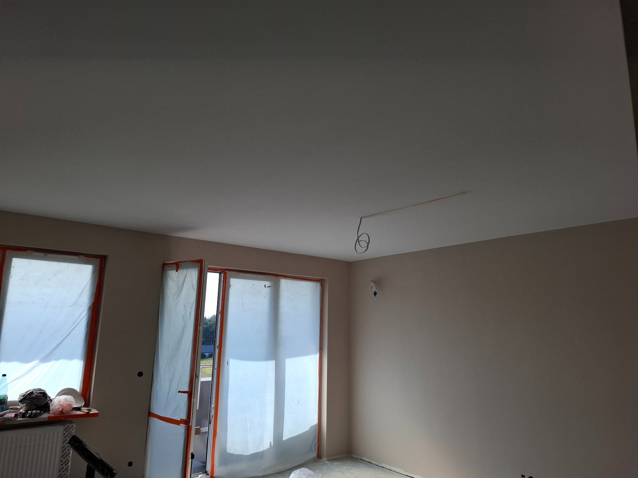 Pokój w trakcie remontu: ściany w neutralnym odcieniu beżu, okna i drzwi balkonowe zabezpieczone folią malarską, widoczne przewody elektryczne na suficie, podłoga częściowo zakryta folią ochronną.