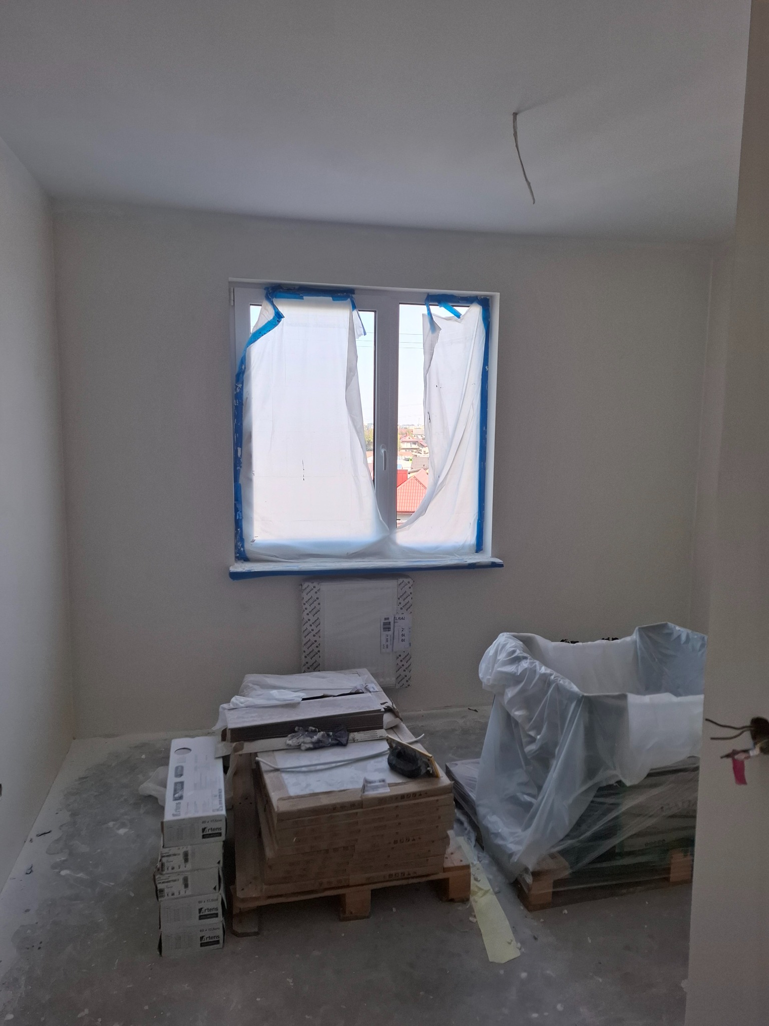 Pokój w trakcie remontu, z zabezpieczonym oknem folią malarską, widoczne materiały budowlane na paletach, ślady po pracach na podłodze, kabel wystający z sufitu.