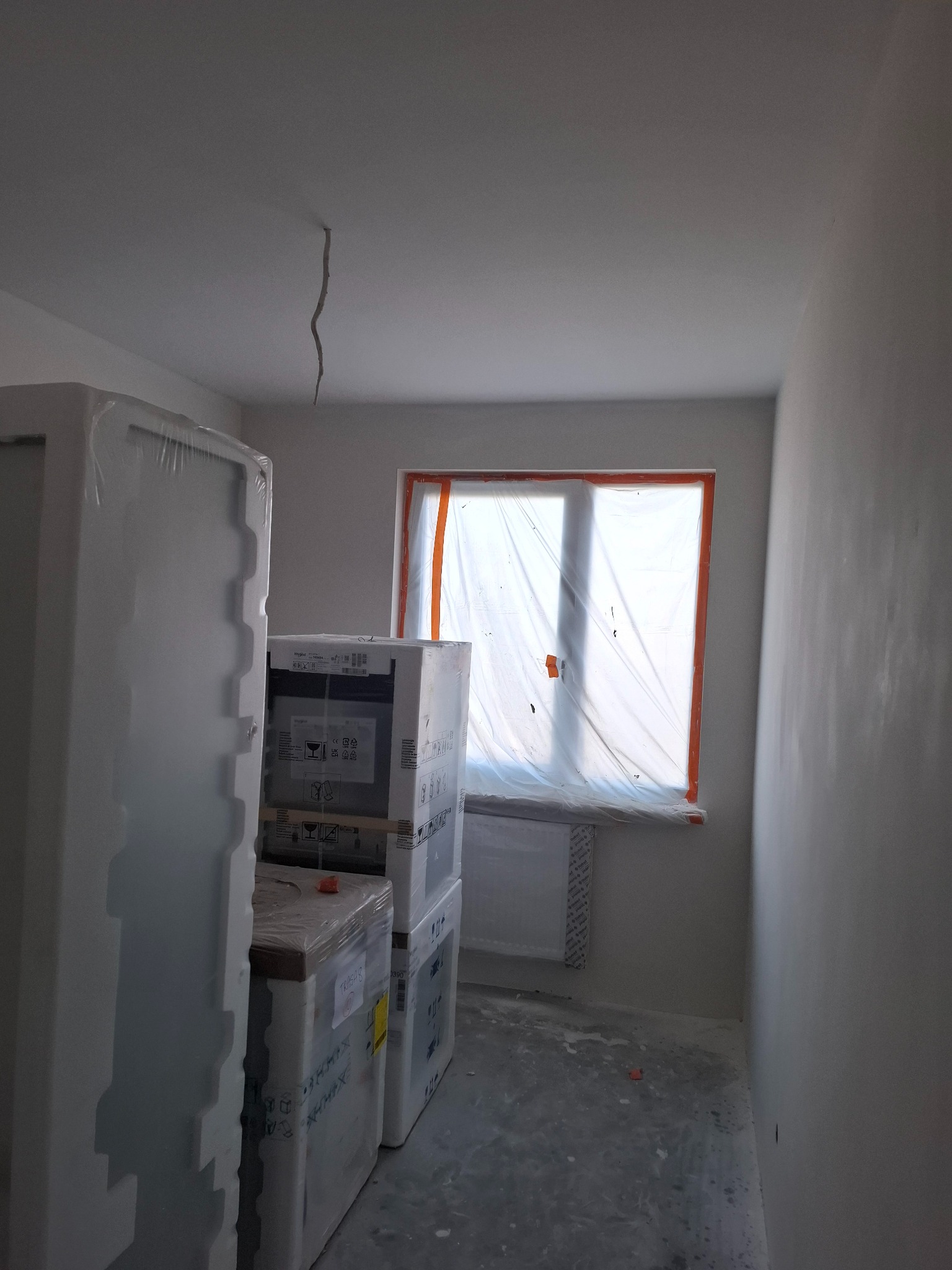 Pokój w trakcie prac wykończeniowych: ściany po nałożeniu gładzi, okno zabezpieczone folią, podłoga z resztkami zaprawy, w rogu zapakowane w folie meble i kartony z armaturą.