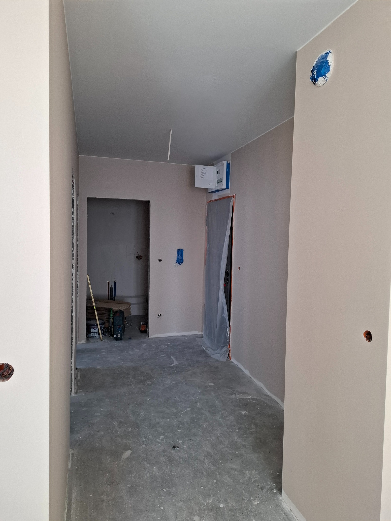 Korytarz w trakcie remontu, ściany przygotowane pod malowanie, widoczne puszki elektryczne, podłoga zabezpieczona, drzwi osłonięte folią malarską, w tle narzędzia i materiały budowlane.