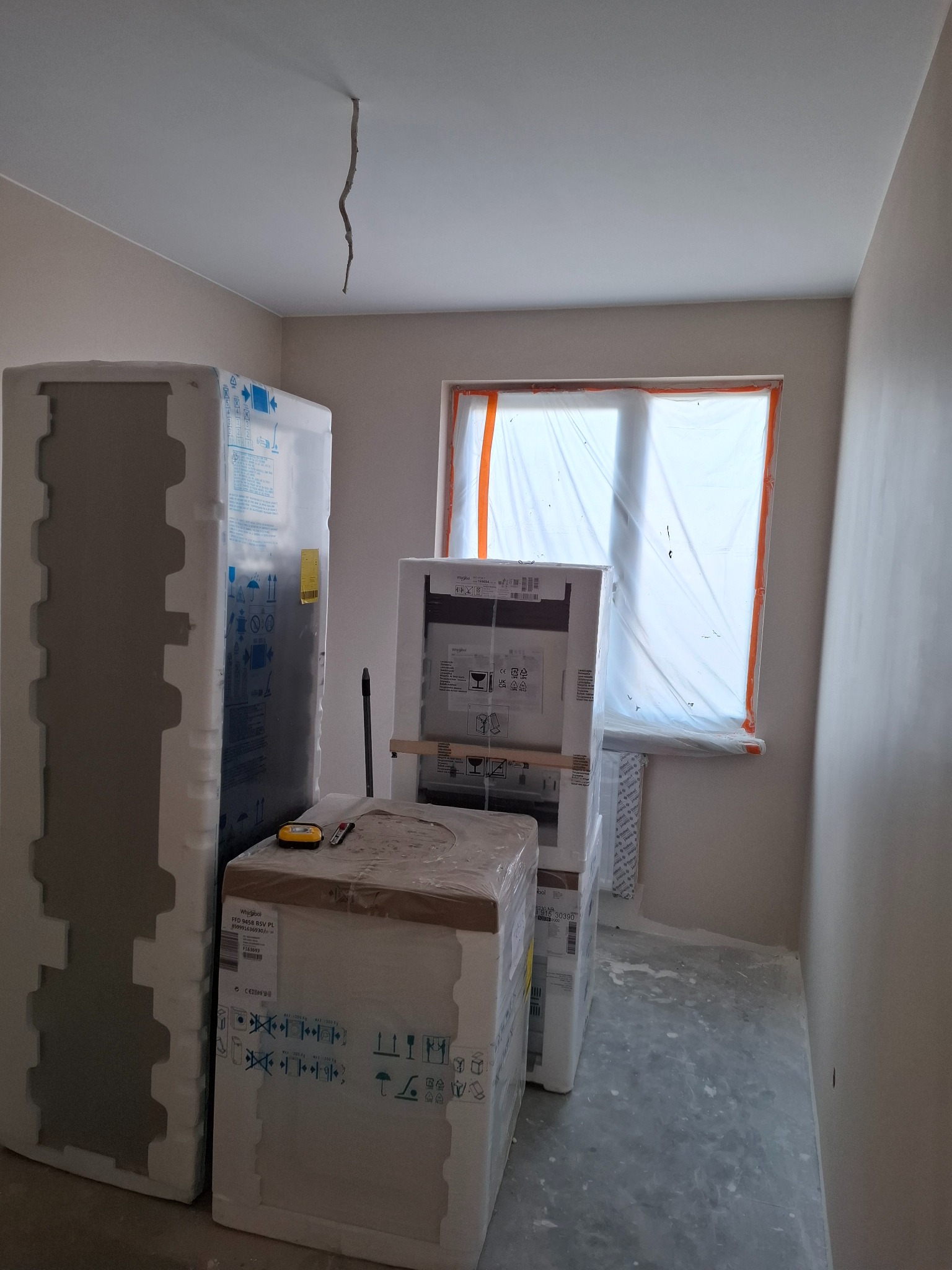 Pokój w trakcie remontu; okno zabezpieczone folią malarską, pod ścianą ustawione w kartonach nowe sprzęty AGD, widoczny fragment niedokończonej instalacji elektrycznej na suficie.
